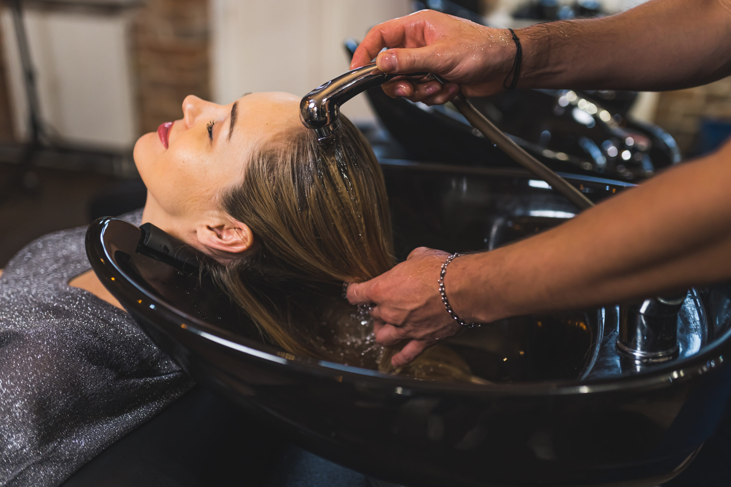 A woman reclining at a salon sink with her head being gently washed by a haircare stylist using a handheld spray nozzle. The setting is a professional hair salon with a relaxing ambiance, and the woman appears to be enjoying the experience.