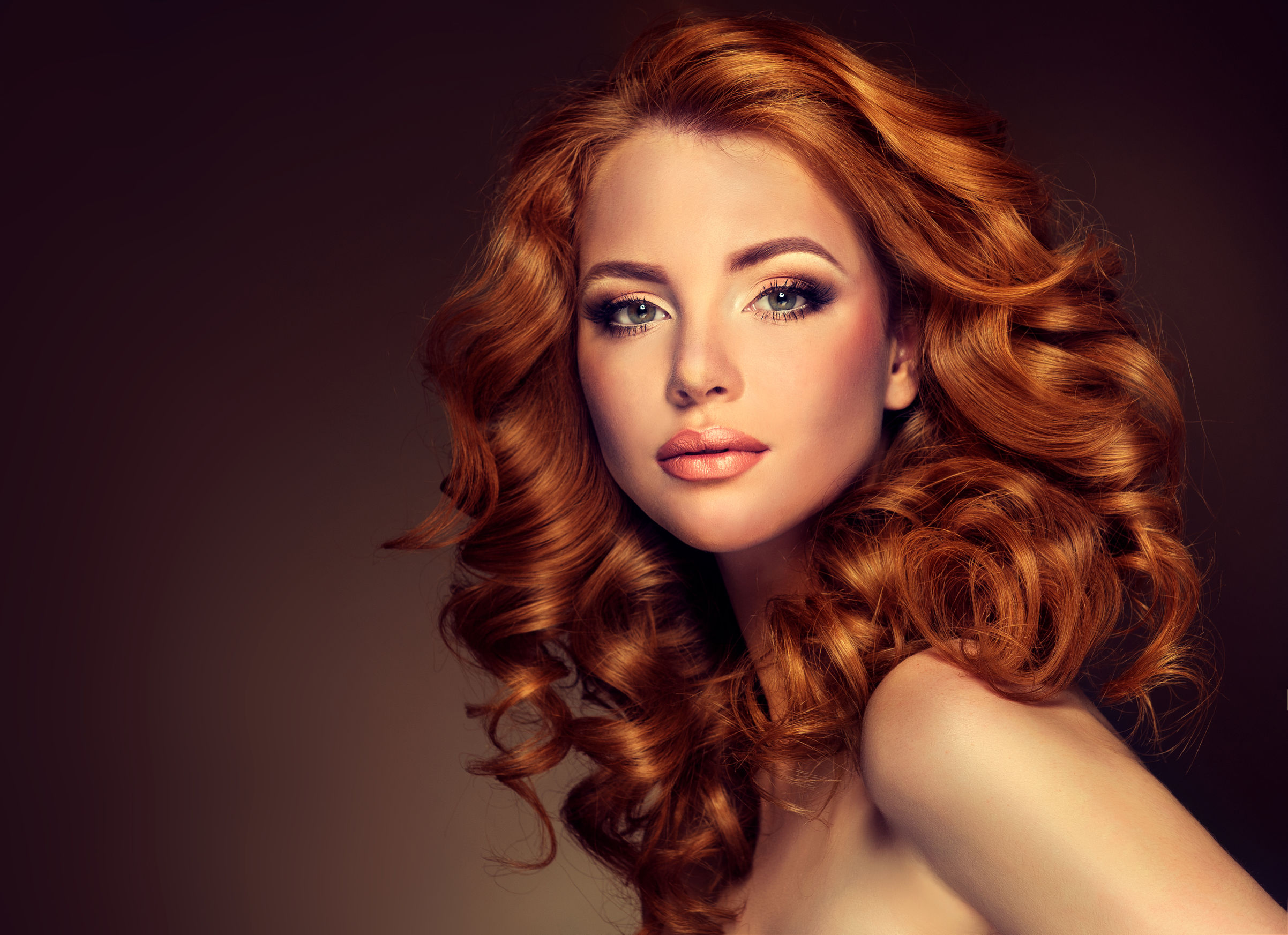 A striking portrait of a woman with vibrant, voluminous red curls cascading around her face and shoulders. Her makeup is elegantly applied, accentuating her eyes and lips, while the warm background complements her hair color, creating a glamorous and sophisticated look.