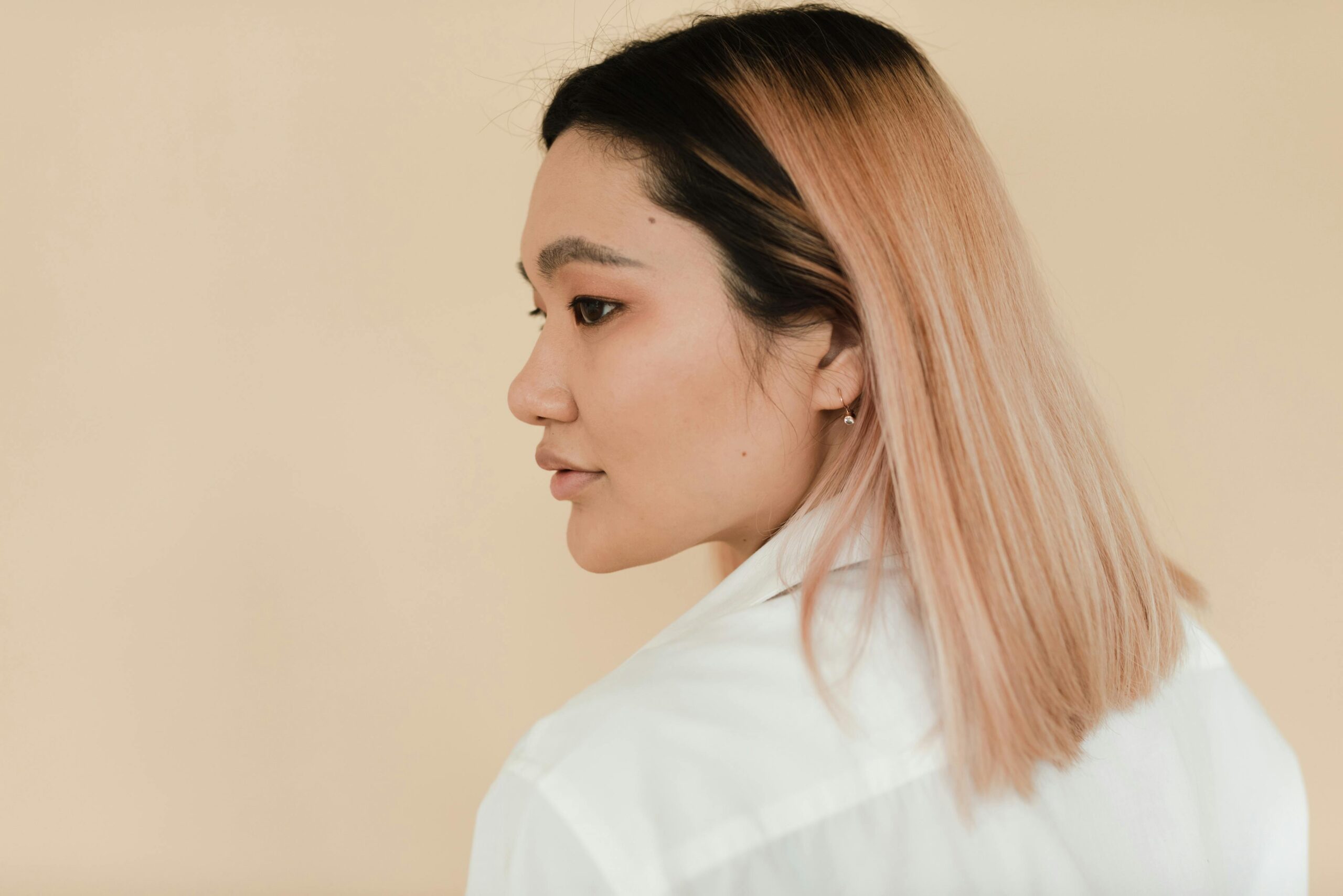 Profile view of a woman with straight, light pink hair wearing a white shirt, looking to her right against a beige background.