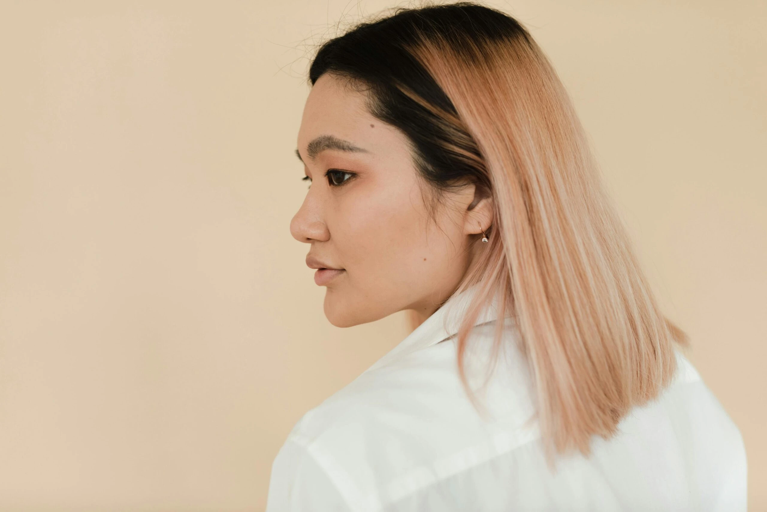 Profile view of a woman with straight, light pink hair wearing a white shirt, looking to her right against a beige background.